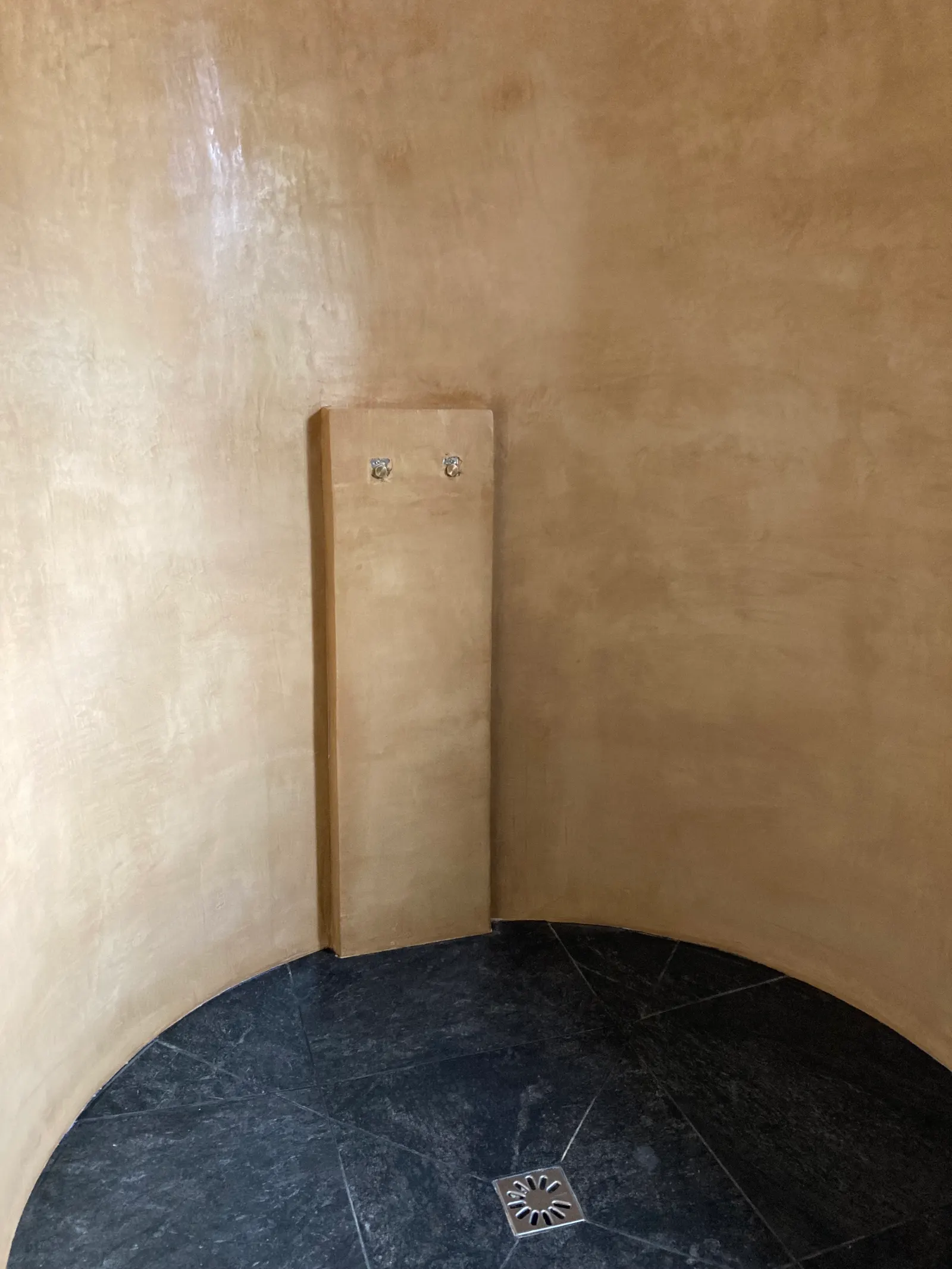 Douche aux murs arrondis en béton ciré beige doré, avec un panneau de robinetterie sur un sol en ardoise