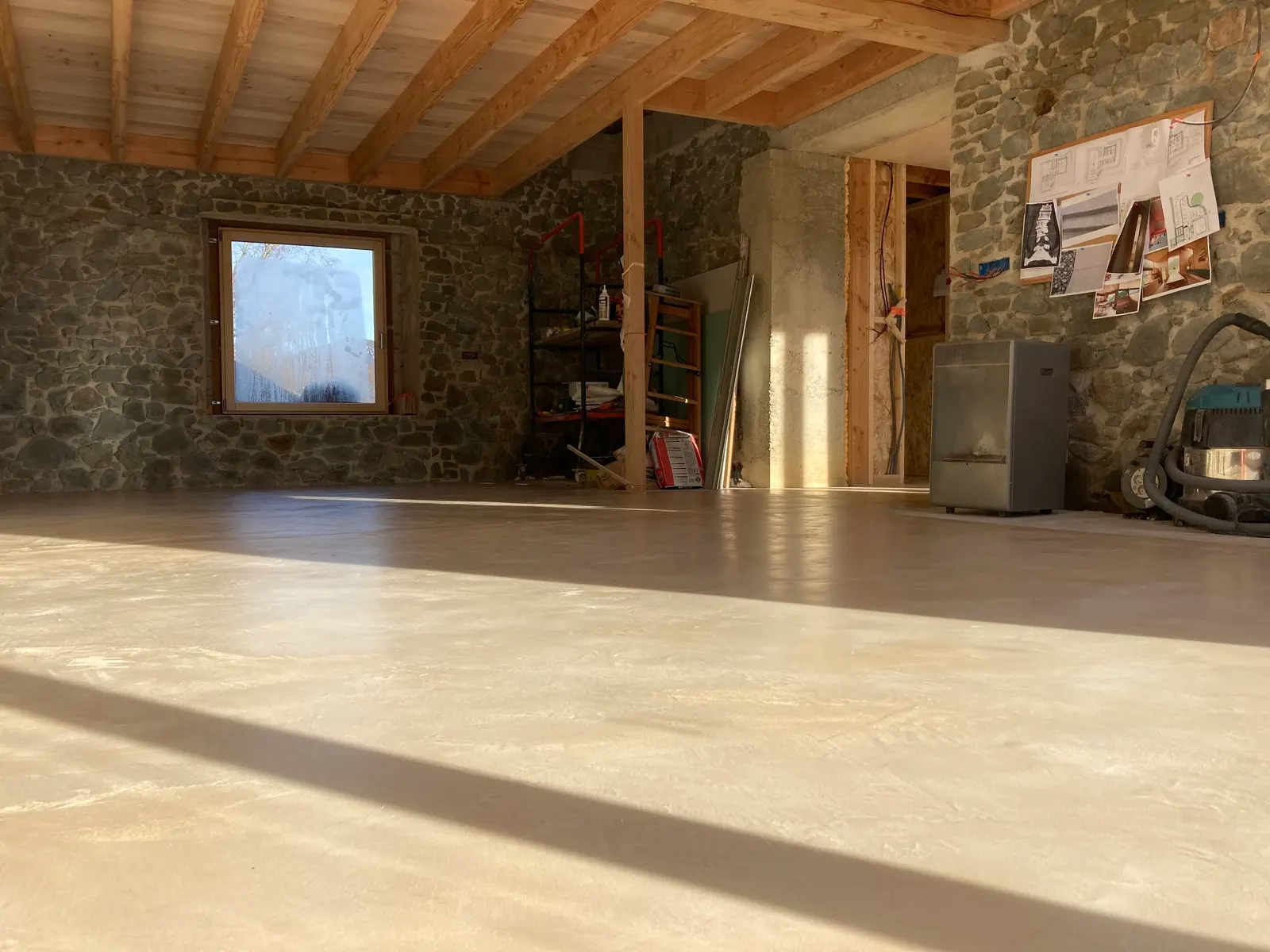 Sol en béton ciré clair dans une grande pièce en rénovation, avec murs en pierre sombre et plafond en bois