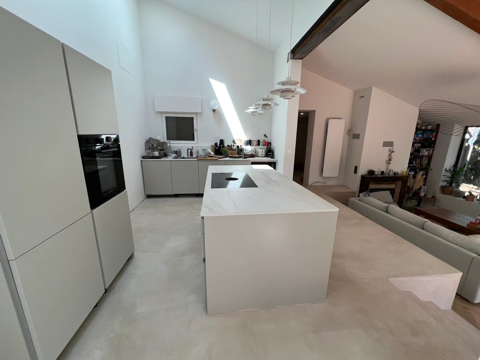 Cuisine blanche avec îlot central et grandes colonnes, sur un sol en béton ciré clair qui se prolonge vers le salon
