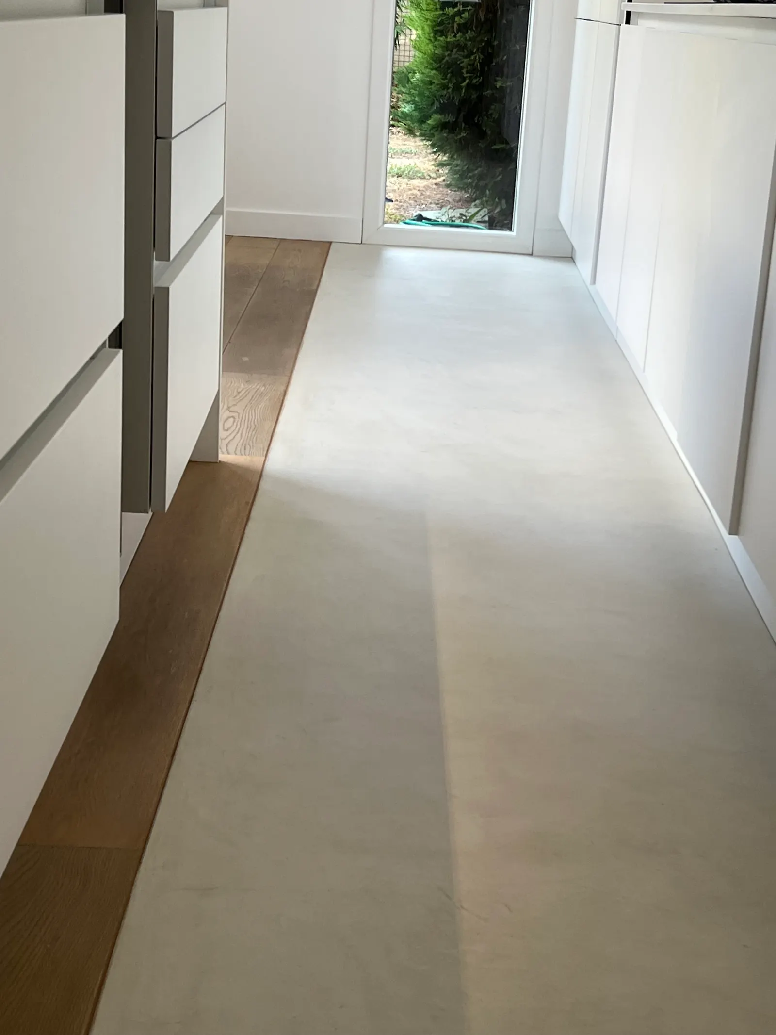 Bande de sol en béton ciré blanc qui longe les meubles de cuisine blancs, bordée par un parquet en chêne