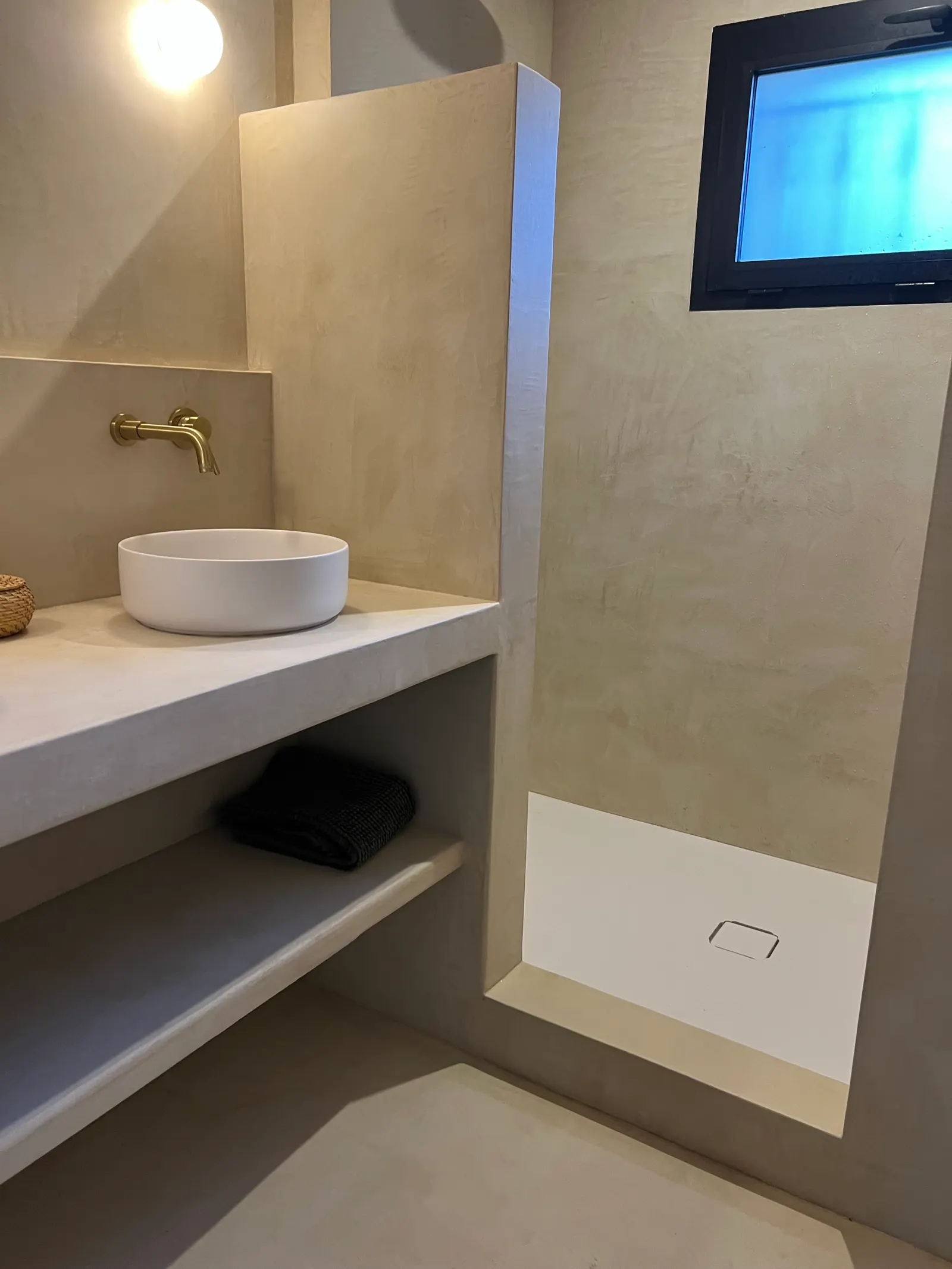 Meuble vasque maçonné en béton ciré sable avec niche ouverte, à côté d'une douche à receveur blanc encadrée de béton ciré