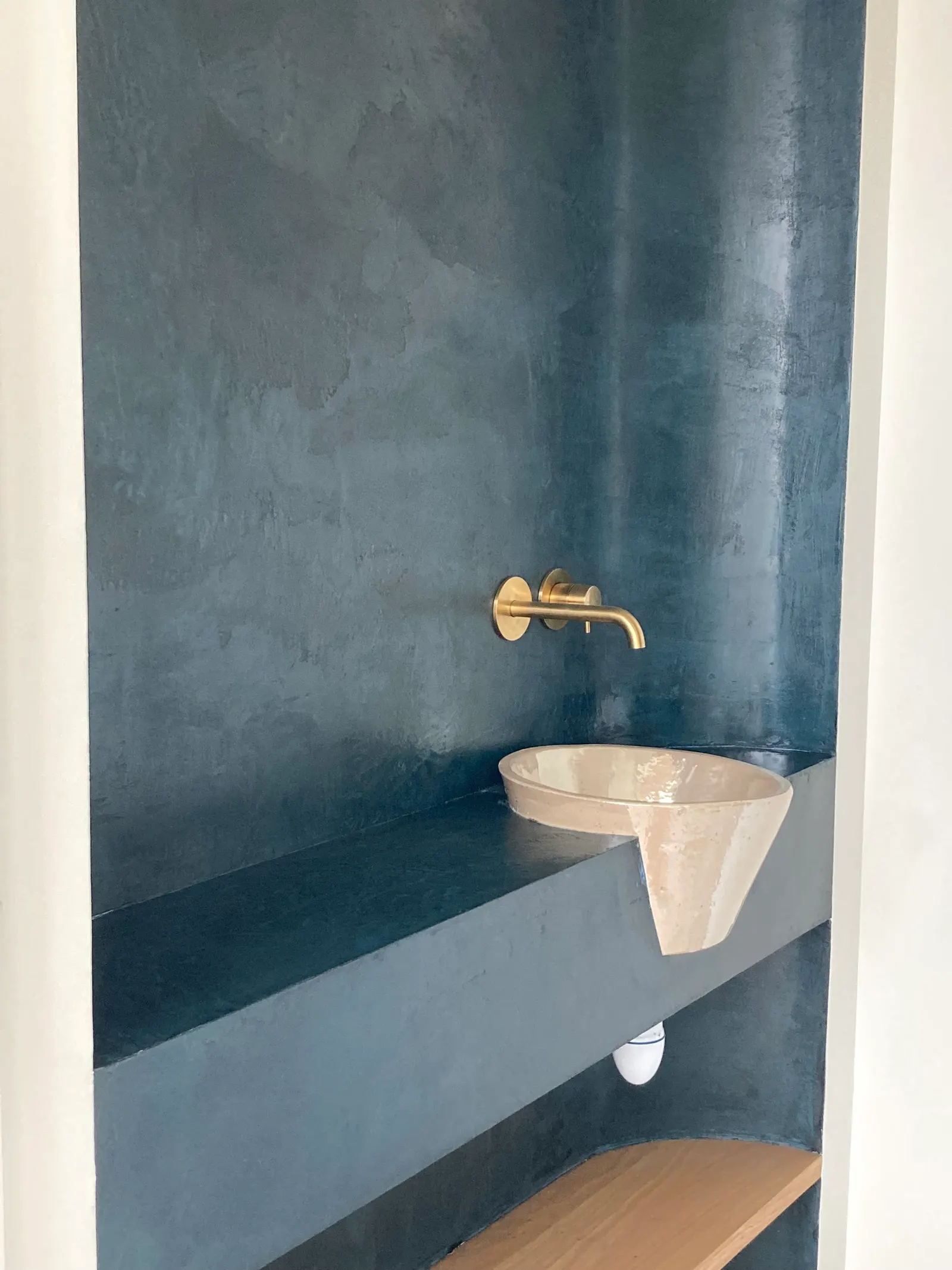 Vue de biais de l'alcôve bleue : plan en béton ciré, vasque conique en saillie et étagère en bois en partie basse