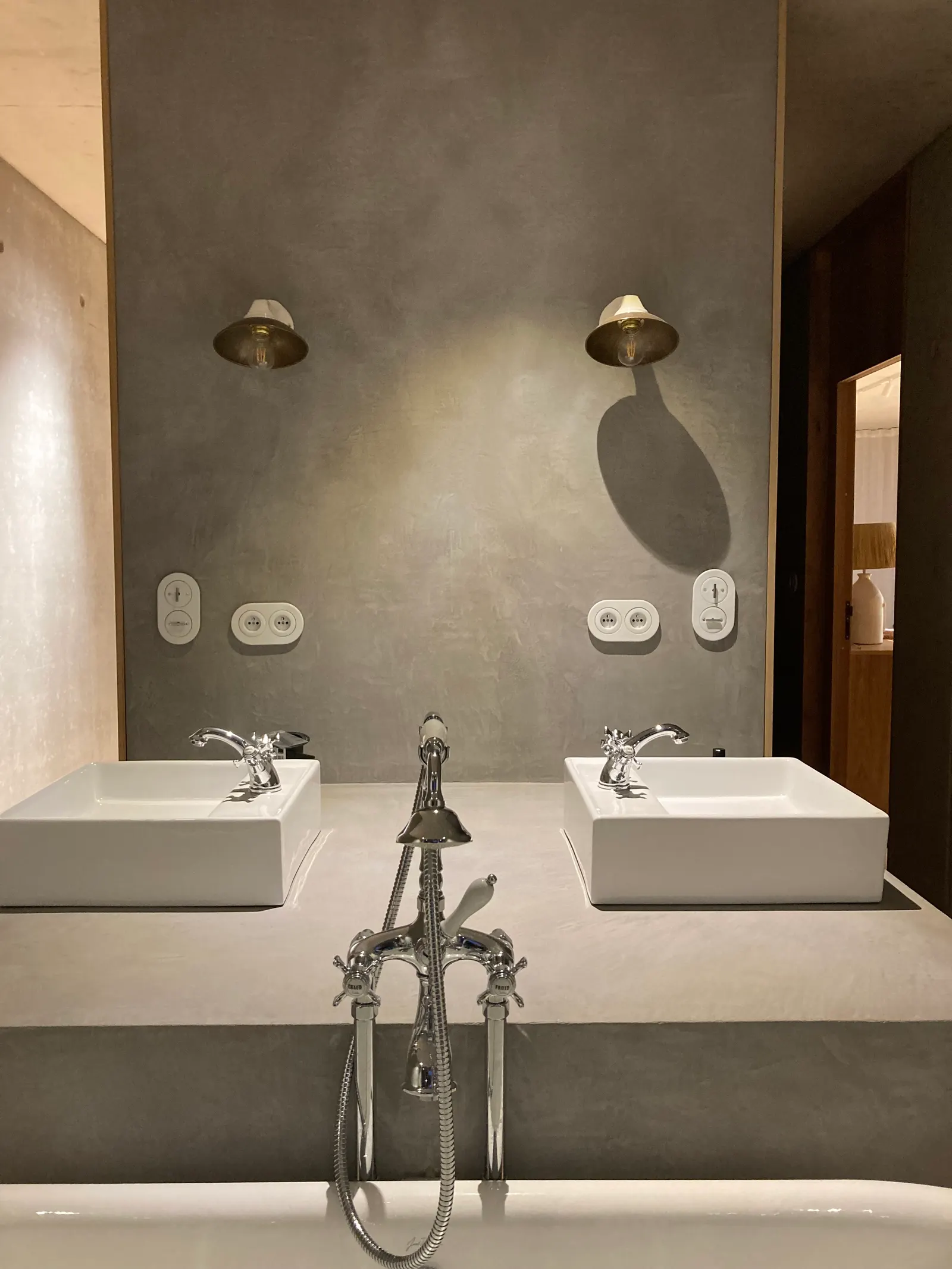 Mur en béton ciré gris nuancé derrière un plan de double vasque assorti, avec appliques en laiton