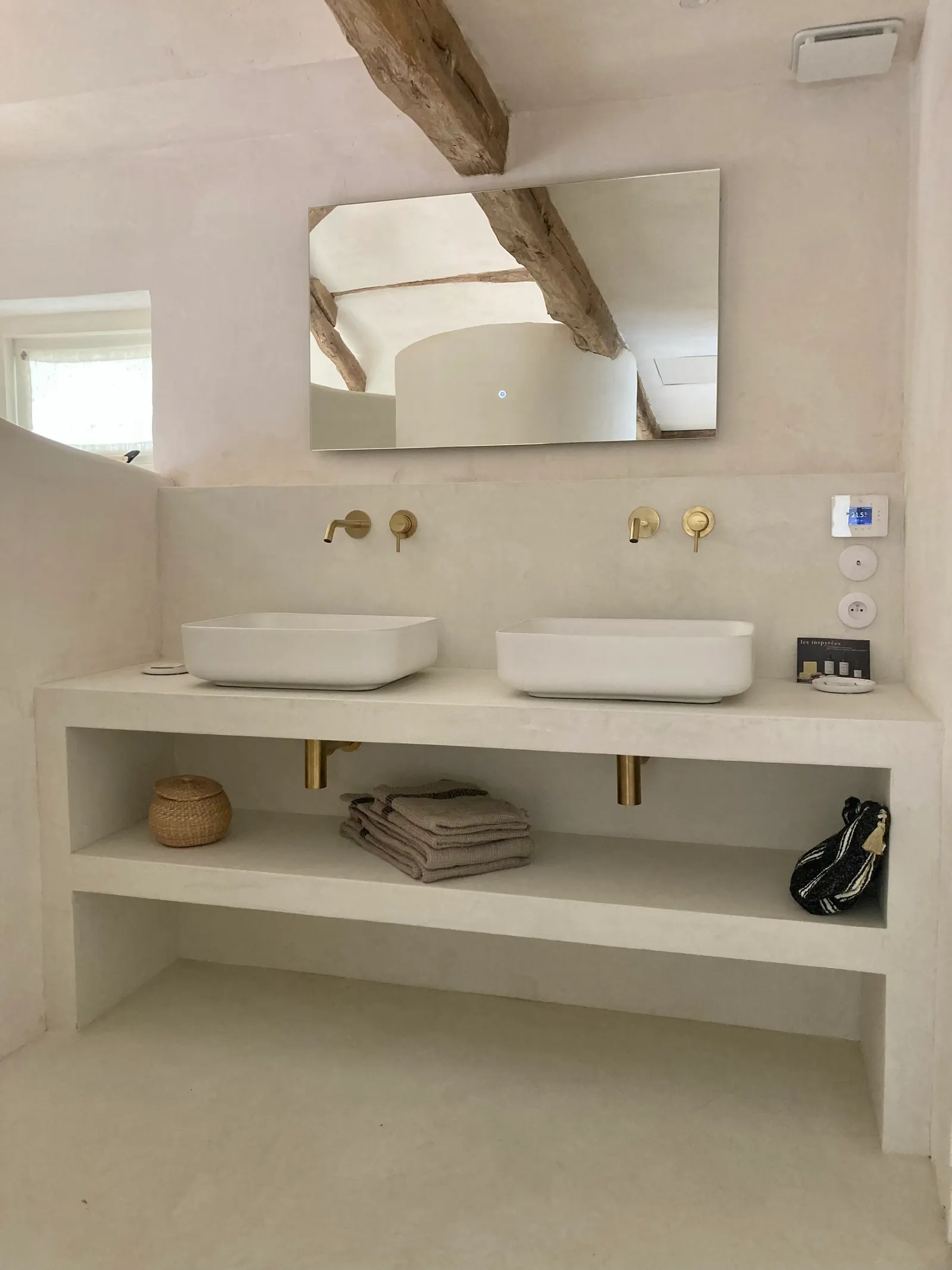 Meuble double vasque maçonné en béton ciré blanc cassé avec étagère ouverte, robinetterie dorée et poutres apparentes