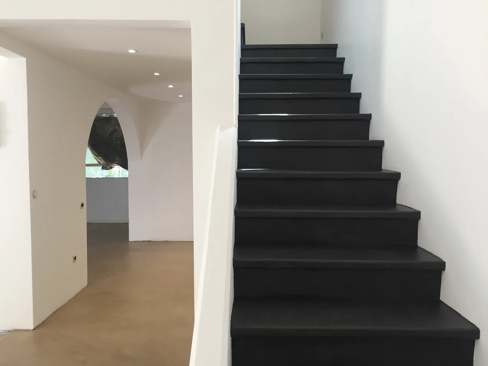 Escalier droit en béton ciré noir entre deux murs blancs, avec un sol de teinte caramel dans la pièce voisine